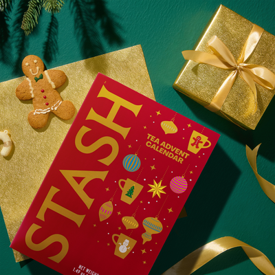 A red Christmas-themed tea advent calendar titled 'STASH' is displayed in a styled flat-lay arrangement on a teal background, surrounded by gold wrapping paper, a gold ribbon, a gingerbread man cookie, and pine branches.