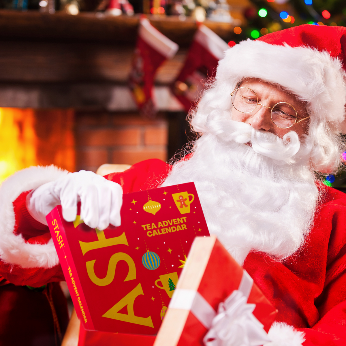 A man dressed as Santa Claus sits in front of a warm fireplace, holding a red 'Tea Advent Calendar' book and a wrapped gift. The background shows stockings and blurred holiday lights.