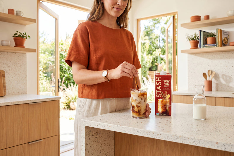 A woman in a rust-orange top stands in a bright kitchen, stirring a glass of iced coffee with a Stash coffee cup on the counter next to a glass of milk.