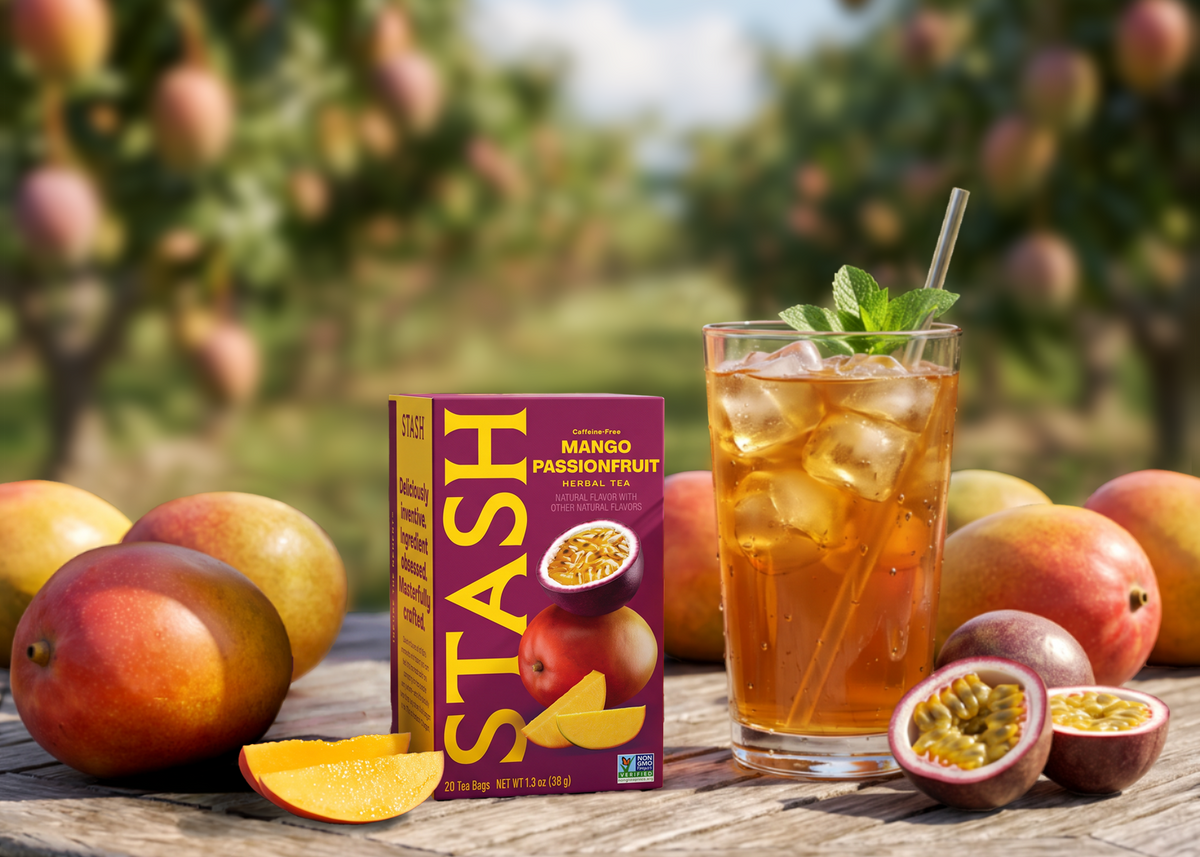 A box of STASH Mango Passionfruit herbal tea stands next to a glass of iced tea with ice and a mint garnish, surrounded by fresh mangoes and passionfruit halves on a rustic wooden table with a blurred passionfruit grove in the background.