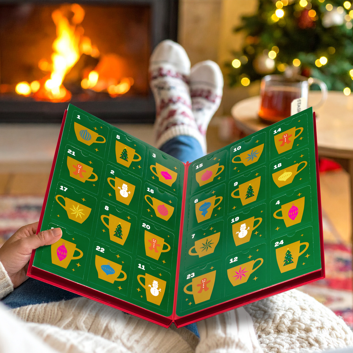 A person sitting by a fireplace with their legs crossed, holding open a Christmas advent calendar made of book pages. The pages are green with numbered yellow teacups and small festive illustrations, while the person wears cozy knit socks and a sweater.
