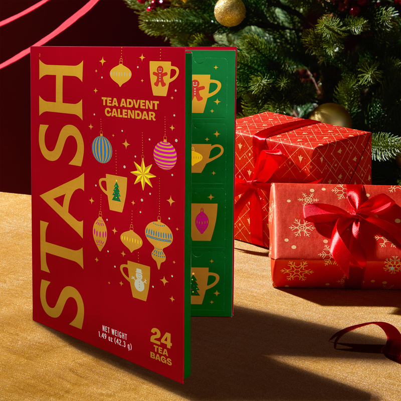 A red and green Stash Tea Advent Calendar box stands open on a tabletop beside wrapped Christmas presents and a decorated Christmas tree. The front of the calendar is red with large gold 'STASH' lettering and holiday illustrations, and the side panel is green with matching illustrations.