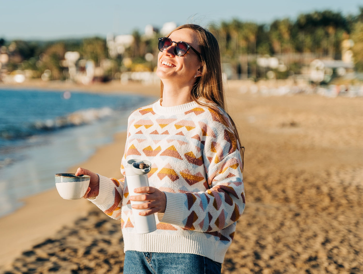 Woman in patterned sweater holding a tea cup and thermos on a beach