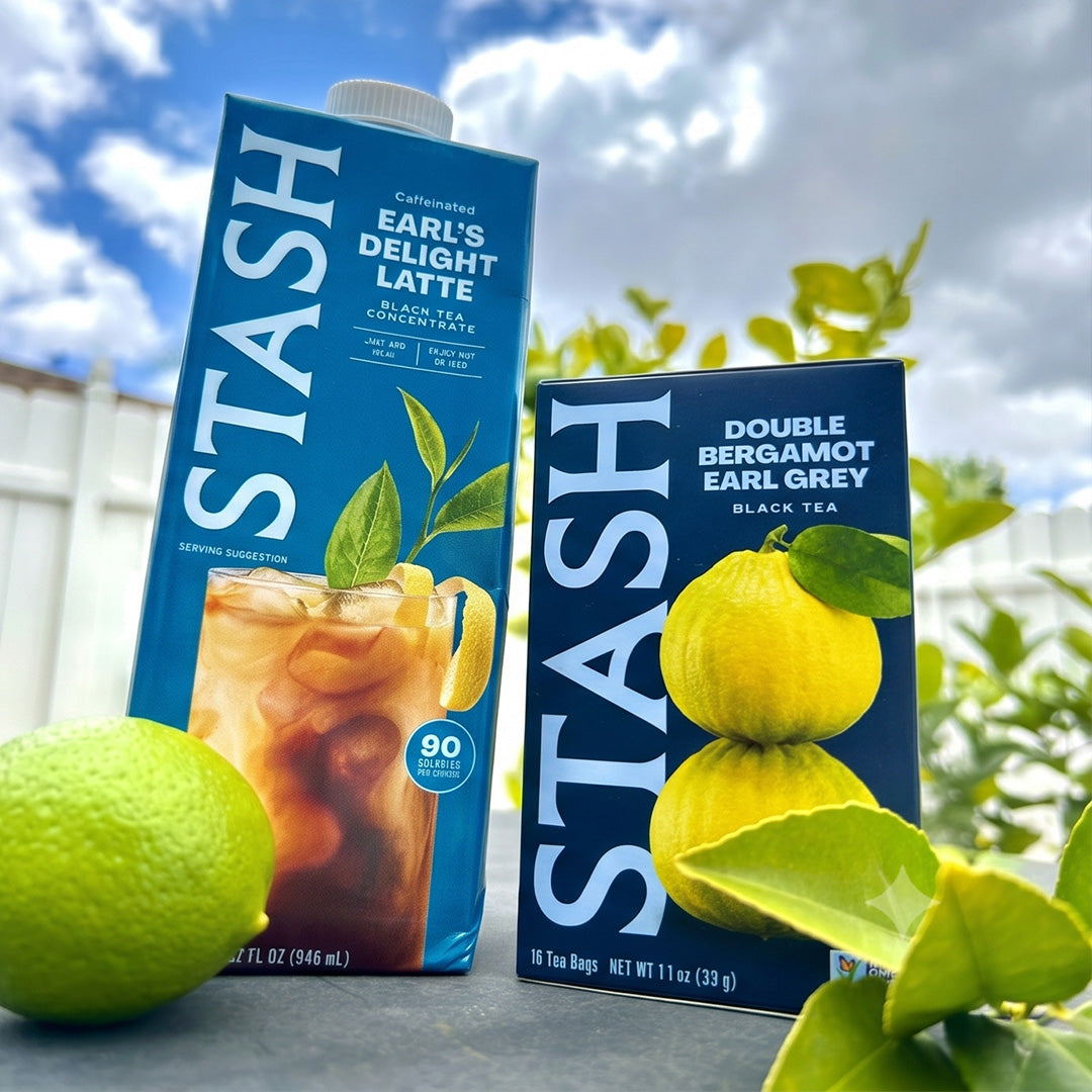 Two Stash tea bags boxes standing outdoors on a table with lemons in the foreground: a tall blue carton of Earl's Delight Latte and a shorter blue box of Double Bergamot Earl Grey. The background shows a white fence, green leaves, and a blue sky with clouds.